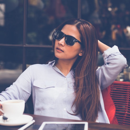 Photo of a young woman wearing sunglasses and a button-up shirt, sitting at a table in a cafe holding a coffee cup