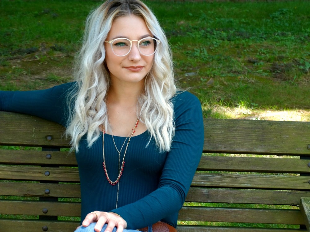 Student wears large, round cream-colored glasses with her long-sleeve blue top and layered jewelry.