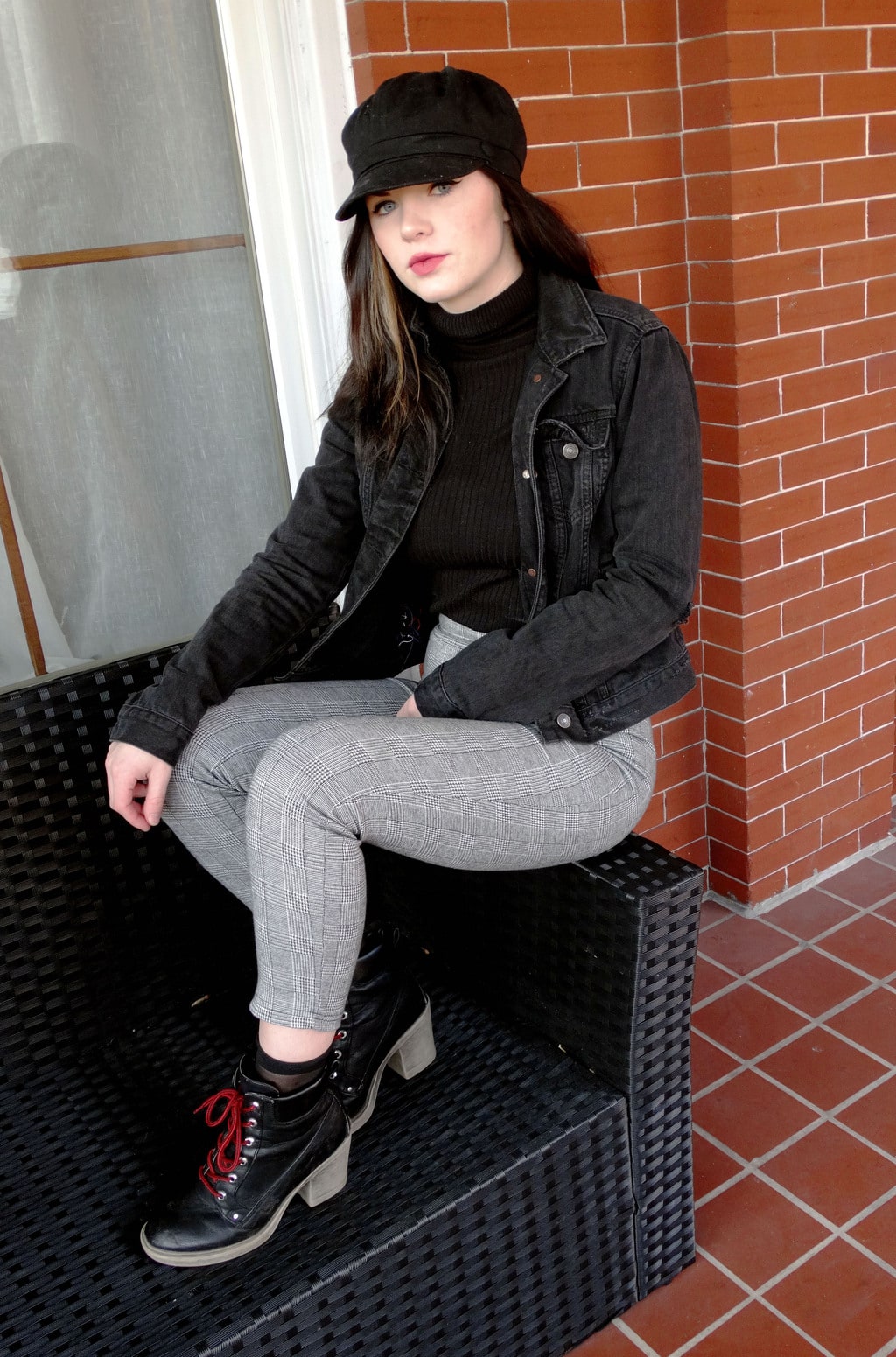 West Virginia University student wears a black turtleneck sweater with white and grey checkered pants, lace up chunky-heeled booties, a distressed dark grey denim jacket, and a black cabby hat.