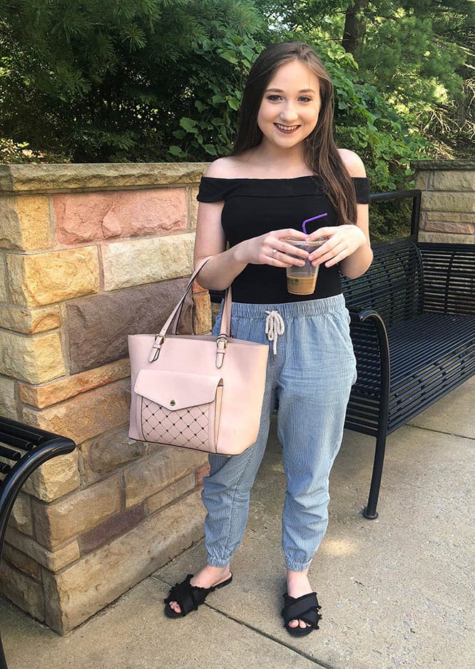 West Virginia University student Maggie wears a black off-the-shoulder fitted top tucked into blue and white drawstring jogger pants with black frayed slide sandals and a pink Michael Kors tote.