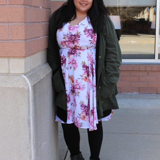 Katie wears a floral print sundress with an olive green jacket, black tights, and grey chunky-heeled lace-up booties.