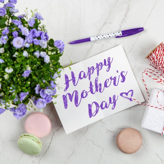 Flat lay on a pale gray marbled surface featuring purple flower plant, uncapped purple-and-white pen with a dotted design; white card that says "Happy Mother's Day" in purple script writing with a heart; a spool of red-and-white string; a small gift box wrapped in white polka-dotted paper and tied with the same kind of red-and-white-string as is on the spool; and three macarons in pale pink, pale green, and light brown colors scattered about