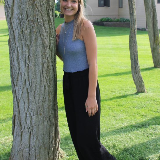 Emily wears a grey high-neck sleeveless tank top, flowy black pants, and layered gold necklaces.