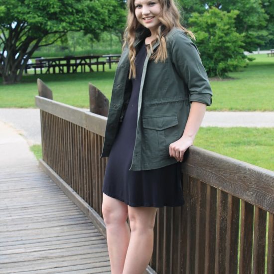 Rebecca wears an army green anorak jacket, a simple black shift dress, and brown criss-cross flat sandals.