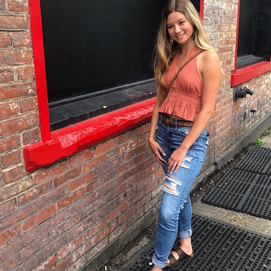 Logan wears a burnt orange halter top with a ruffle peplum, ripped and cuffed skinny jeans, and dark brown birkenstock sandals.