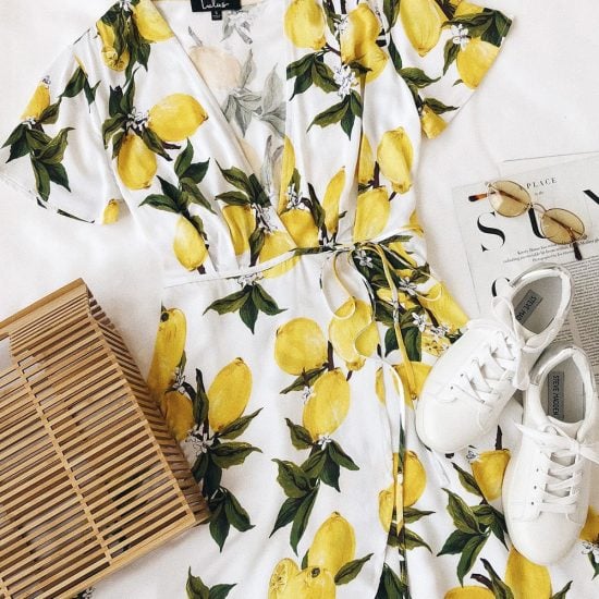 Flat lay featuring short-sleeved lemon print wrap dress, wooden bag, small oval sunglasses, and white sneakers, with part of an open magazine visible in the background