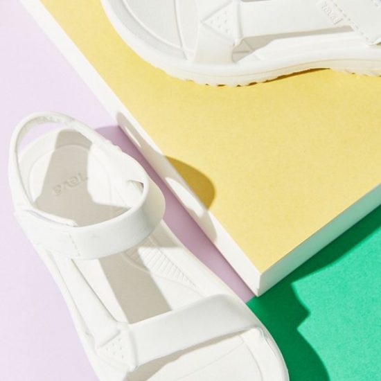 White rubber Teva sandals against a green and pastel yellow and lilac-colored background.