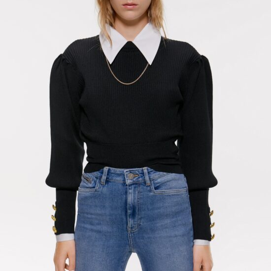 Young woman wearing a black puff-sleeve sweater with three gold buttons at each cuff over a white shirt with an oversized collar, as well as a skinny gold chain necklace and light-wash skinny jeans