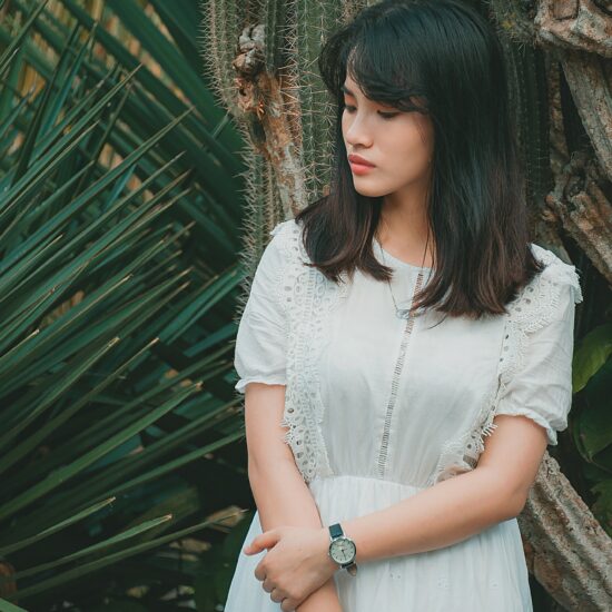 Woman wearing short-sleeved white dress with lace details and a watch with a black strap standing against a background of large plants and cacti