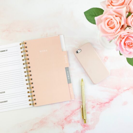 Flat lay on light pink marble surface featuring planner, gold-colored pen, iPhone in pale pink case, and pale pink roses