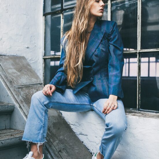 Woman with very long blond hair sitting on a window ledge outside next to stairs, looking away from the camera and wearing a blue plaid double-breasted blazer, loose-fitting light-wash jeans with distressed hems, and Adidas sneakers