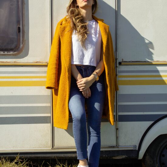 Woman standing outside in front of vehicle wearing yellow coat over white boxy-fit t-shirt and skinny jeans, as well as a watch and a bracelet, earrings, cat-eye sunglasses, and white sneakers