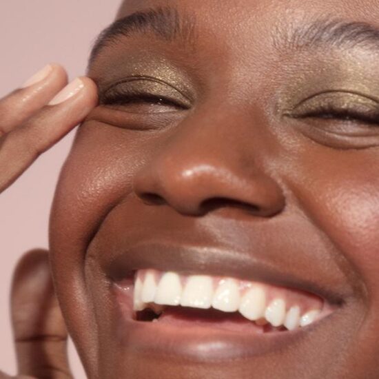 Close-up shot of woman smiling against pale pink background and wearing Glossier Lidstar in "Herb." She is gently touching the corner of her right eye with the finger of her right hand.