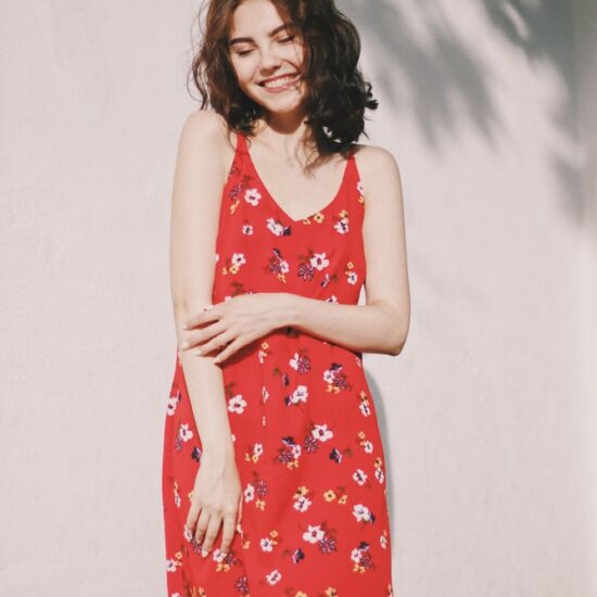 Smiling young woman wearing sleeveless red floral-print dress standing against white wall outside