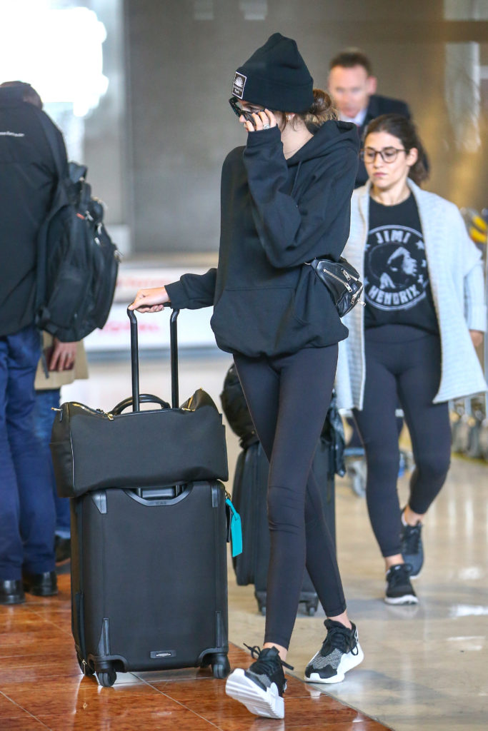 Kaia Gerber simple airport outfit with black leggings, black and white sneakers, oversized black hoodie, fanny pack and beanie